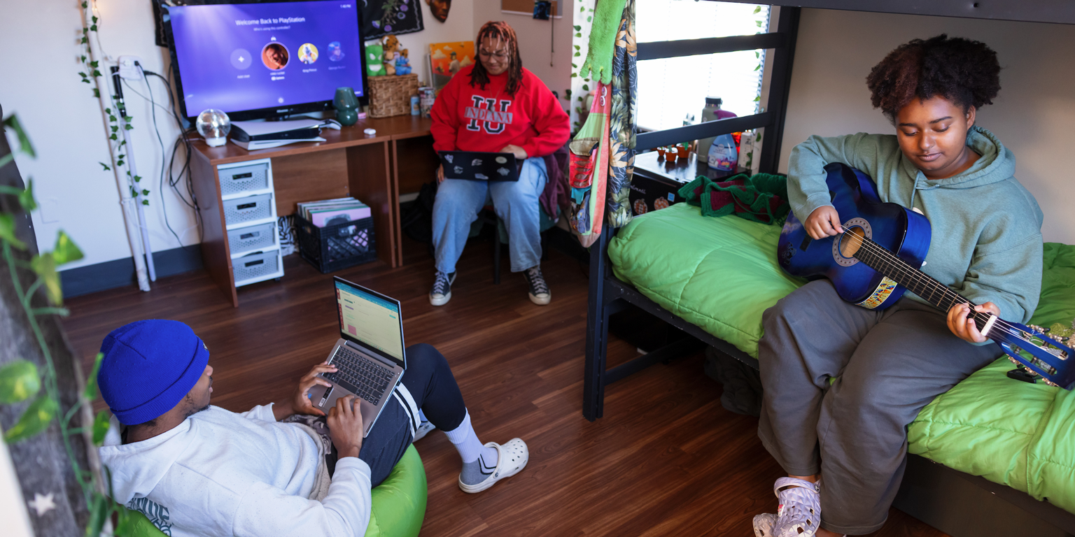 Two female students in a dorm room. One is working on her laptop, and the other is playing an acoustic guitar.